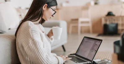 Side view of female freelancer in warm sweater and eyeglasses drinking tea from white ceramic cup while sitting on floor near sofa with netbook on legs while creating document for remote work project